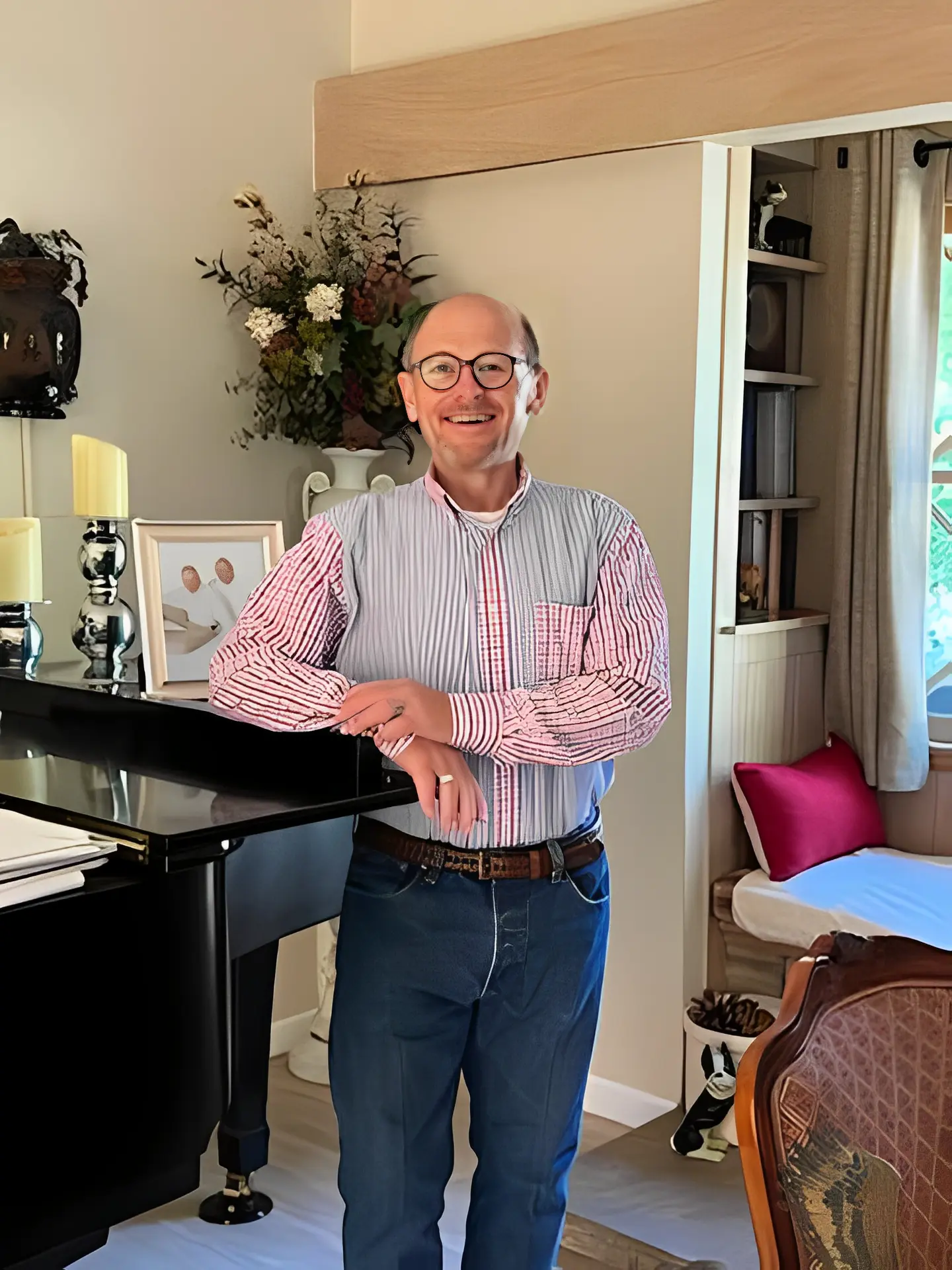 A smiling man leaning on a piano in a cozy room.